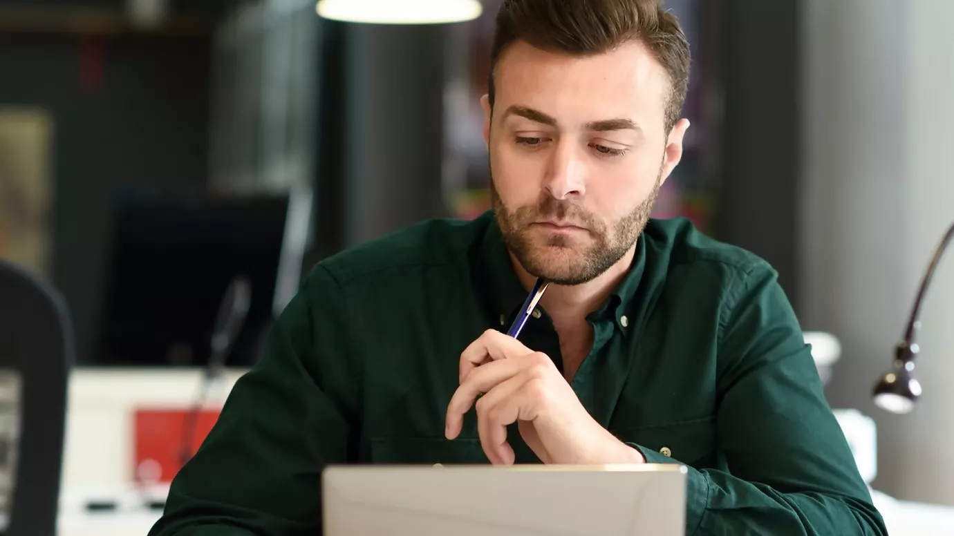 Un homme concentré travaillant sur un ordinateur portable dans un bureau moderne, tenant un stylo en réfléchissant.