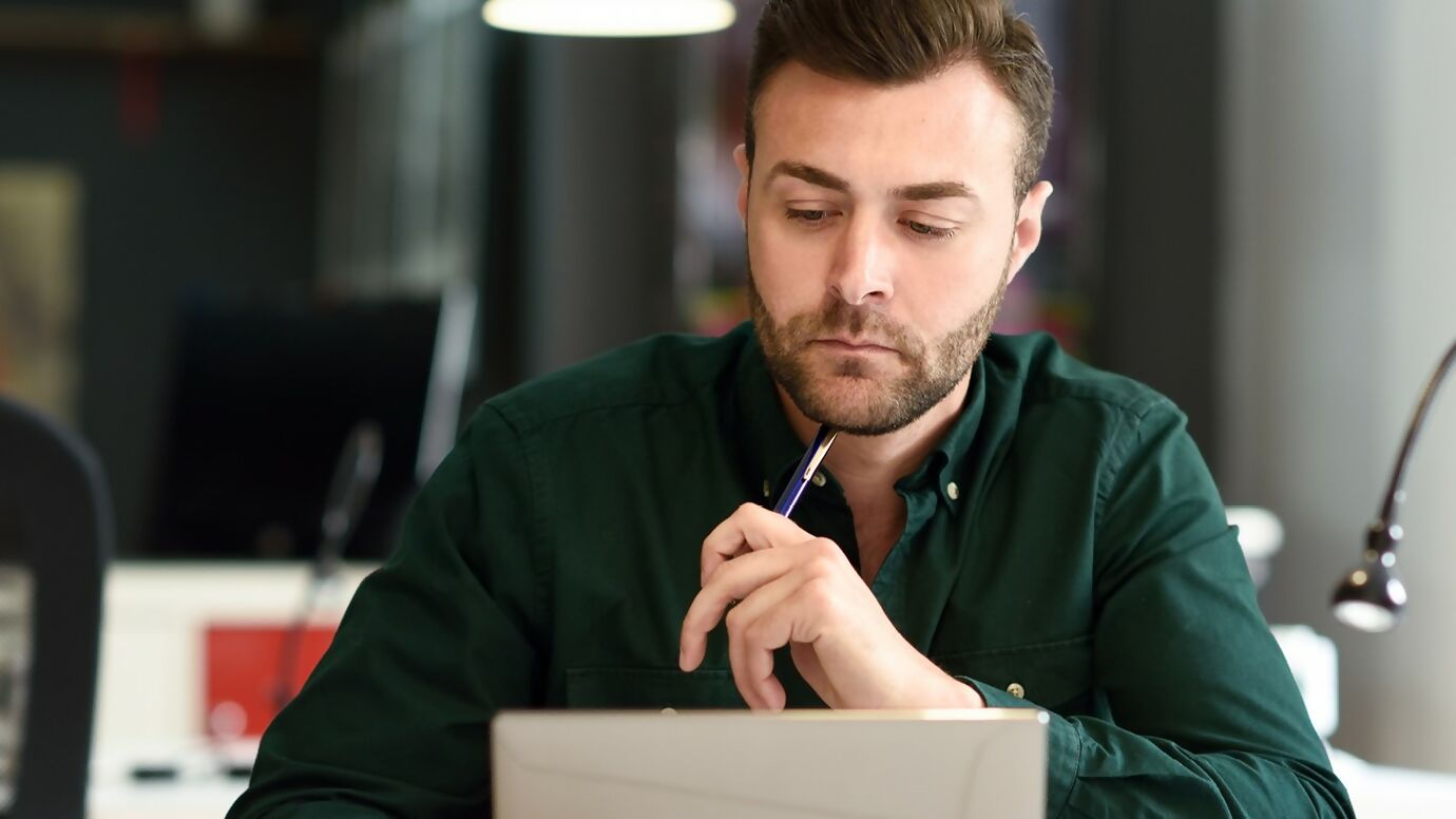 Un homme concentré travaillant sur un ordinateur portable dans un bureau moderne, tenant un stylo en réfléchissant.
